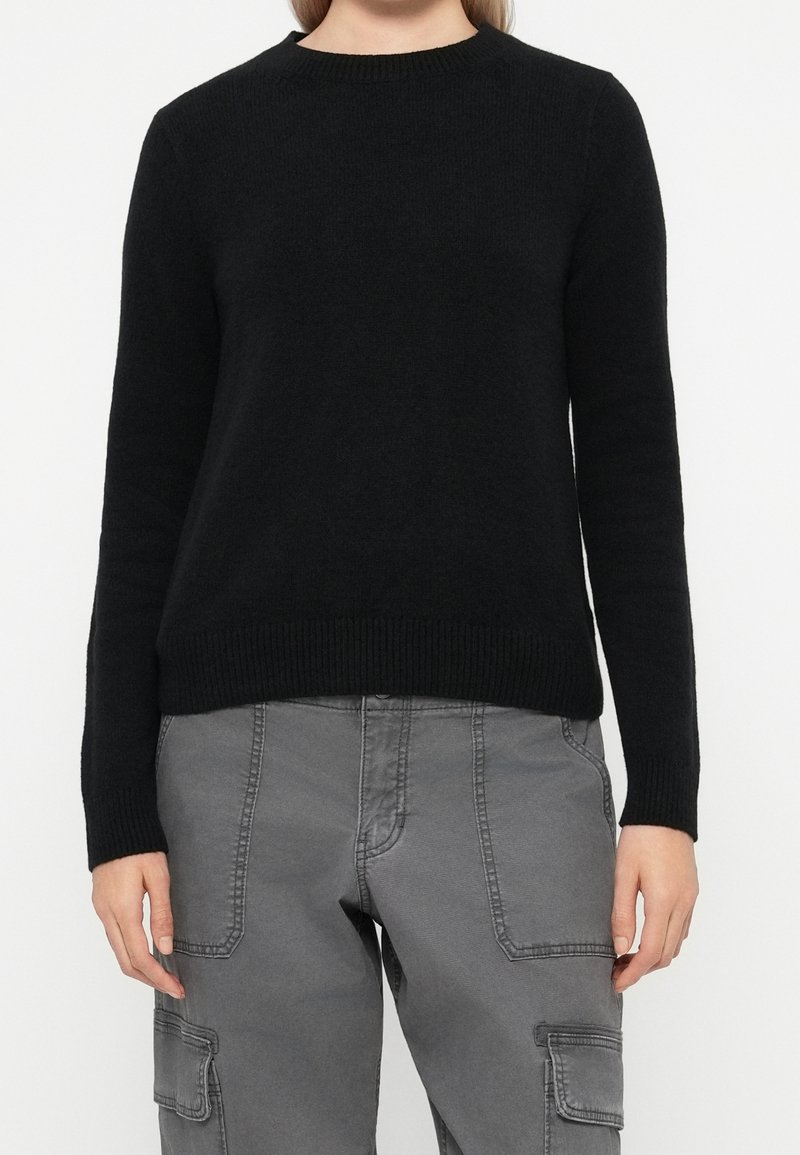 Personne portant un pull en maille noir à manches longues et un pantalon cargo gris avec de grandes poches avant, debout devant un fond blanc uni.