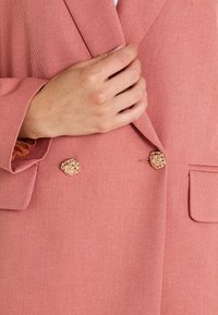 Blazer rose en tissu texturé, doté d'un design croisé avec des boutons décoratifs dorés et une poche poitrine unique.