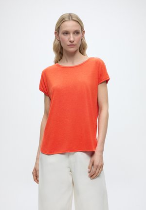 Mujer con cabello claro que lleva una blusa de manga corta naranja brillante y pantalones blancos sueltos, de pie frente a un fondo claro y liso.