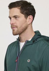 Grüne Kapuzenjacke mit Reißverschluss vorne, rotem Reißverschlussanhänger und Logo-Patch. Der Stoff wirkt glatt mit matter Oberfläche. Darunter ist eine weiße Grundschicht.