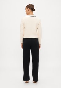 Zilo krekliņu ar melnu apkakli un garām piedurknēm, kas savienots ar melniem piegrieztiem biksēm un bēšām kurpēm ar papēdi, skatīts no aizmugures.