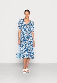 Vestido floral com um padrão azul e branco, decote em V, mangas curtas bufantes, cintura atada, até ao joelho. Combinado com uma bolsa azul clara e sandálias brancas.