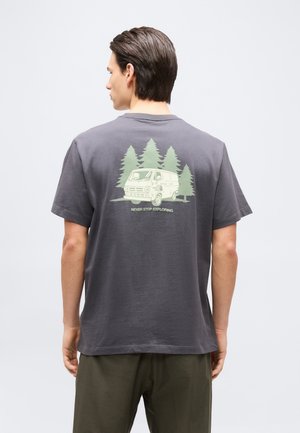 Hombre con camiseta gris oscuro con un gráfico de una furgoneta vintage y árboles de pino, y el texto "Nunca dejes de explorar" en la parte trasera.