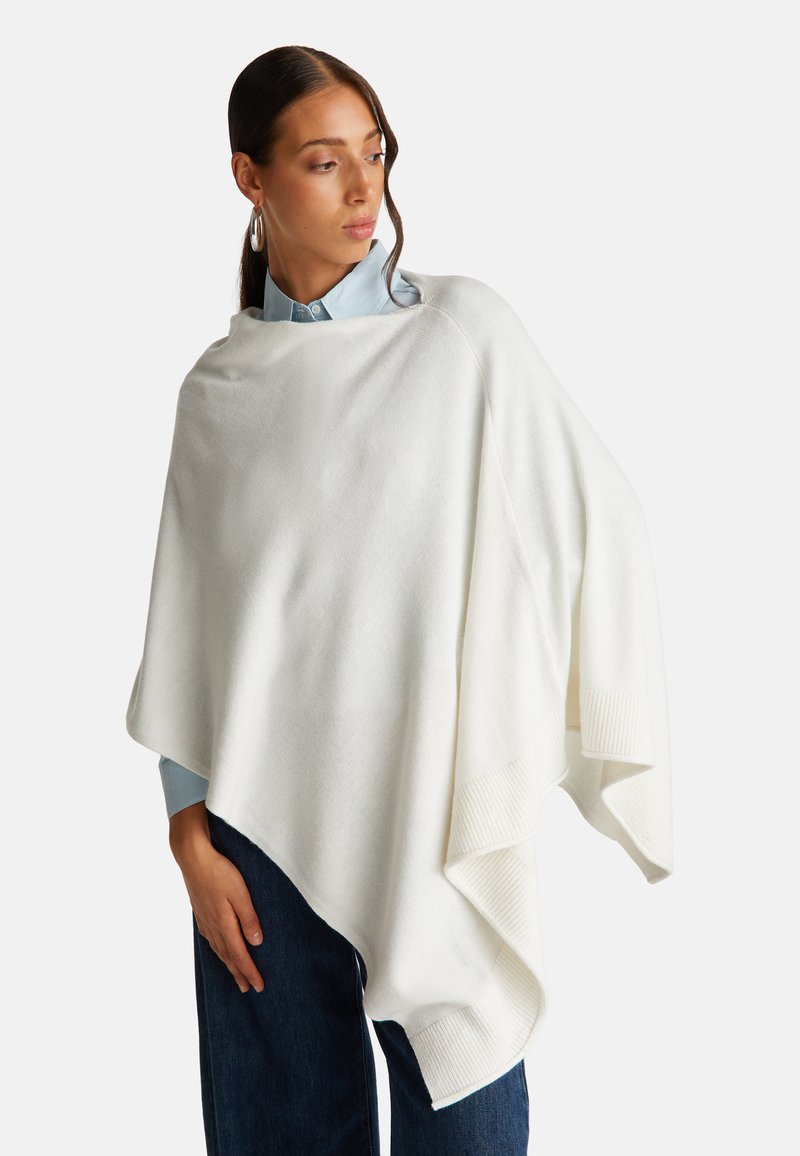 Bílý, oversized pončo vyrobené z měkké látky. Má design s drapérií a žebrovanými okraji, který doplňuje světle modrá košile s límečkem a tmavé džíny.