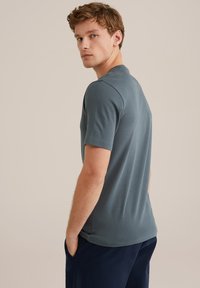 Camiseta de manga corta para hombre en azul pizarra, hecha de un tejido suave, con cuello redondo y ajuste slim. El modelo está de pie con las manos en los bolsillos.