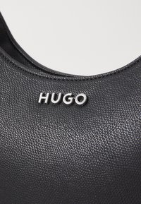 Černý texturovaný syntetický materiál s kulatým výstřihem. Stříbrné logo "HUGO" výrazně vyobrazené v přední části. Viditelné švy.