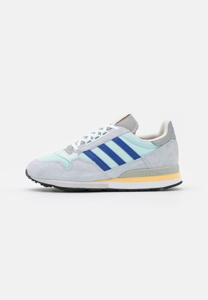 Baskets Adidas bleu clair et gris avec des bandes bleu foncé, des lacets blancs et un accent jaune sur la semelle, vues de côté sur fond blanc.