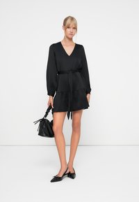 Vestido negro de manga larga con escote en V, cintura ceñida, falda en capas y textura suave. Combinado con un bolso tipo cubo negro y zapatos planos.