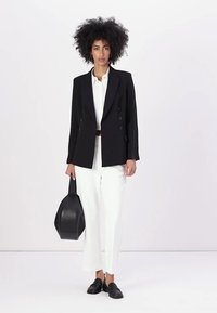 Blazer preto de dupla abotoadura com lapelas largas, combinado com calças brancas. O modelo segura uma mala preta e usa mocassins. Textura suave, ajuste feito à medida.