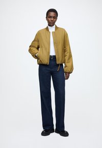 Żółta kurtka typu bomber z zamkiem błyskawicznym, prążkowanym kołnierzem i elastycznymi mankietami, w połączeniu z niebieskimi, szerokimi spodniami jeansowymi i czarnymi, masywnymi butami.