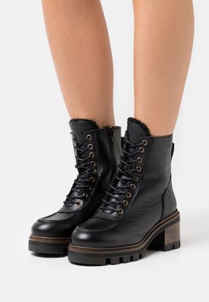 Black leather lace-up ankle boots with chunky heels, side zippers, and fur lining, worn on bare legs against a white background.