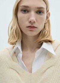 Beige gestrickter Pullover mit V-Ausschnitt, über einem weißen Button-up-Hemd getragen. Weicher Stoff, leicht lockere Passform und Rippdetails am Pullover.