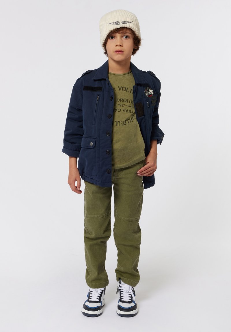 Marine knoopjack, groen graphic t-shirt, olijfgroene broek, witte beanie en blauw-geaccentueerde sneakers gedragen door een rechtop staand kind.