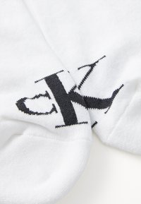 Fehér pamut zoknik, amelyek oldalán feltűnően látható a fekete "CK" logó, puha, sima textúrával és klasszikus dizájnnal.