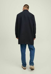 Lungo cappotto blu navy realizzato in tessuto spesso, con colletto classico e taglio dritto, abbinato a pantaloni in denim blu chiaro e scarpe beige.