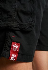 Shorts noirs avec une étiquette en tissu rouge près de l'ourlet affichant "Alpha Industries Inc U.S.A" et le texte "Remove Before Flight".