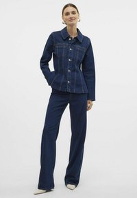 Temno denim jakna z dvema sprednjima žepoma, srebrnimi gumbi in rumenim šivom, v kombinaciji s temnimi kavbojkami in ostro bež obuvalo.
