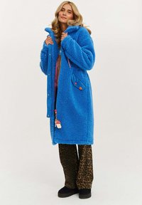 Modrá fleecová bunda s predným zipsom, bočnými vreckami a kapucňou. Nohavice s leopardím vzorom. Hladká textura a živá farba.