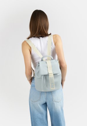 Donna che indossa uno zaino azzurro chiaro con spallacci crema sopra una canottiera bianca e jeans blu, in piedi davanti a uno sfondo bianco uniforme.