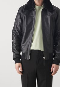 Svart läderbomberjacka med svart pälskrage, dragkedja fram, ribbade ärmslut och en jämn yta. Bärs över en ljusgrön skjorta.