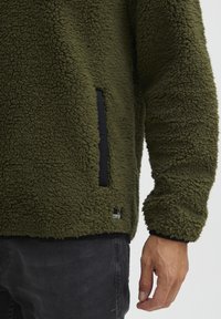 Olive green fleece pullover with a textured surface, featuring a black side pocket and a small logo tag on the bottom left.
