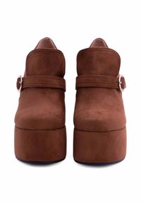 Stivaletti ankle platform in suede marrone con punta tondeggiante, caratterizzati da una fascia e un dettaglio in metallo con fibbia. Texture liscia e design della suola elevata.