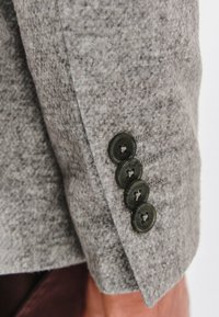 Szary wełniany marynarka z mankietem, ozdobiona czterema ciemnozielonymi guzikami o gładkiej teksturze i subtelnymi detalami szycia. Proste, klasyczne elementy designu.