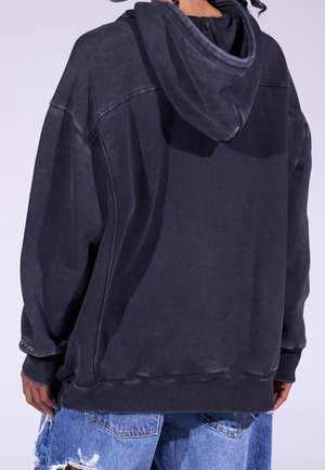 Osoba nosící přehnaně velkou vybledlou černou mikinu s kapucí staženou dozadu a roztrhané modré džínové šortky stojící zády ke kameře.