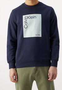 Mörk marinblå sweatshirt med en ljusblå fyrkantig grafik som har texten "Calvin Klein" och "Est. 1968" nedanför, med ribbade mudd och nederkant.
