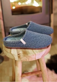 Soft, blue, fleece-textured slippers resting on a round wooden stool with a blurry warm-lit background.