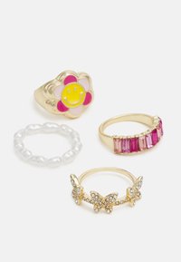 Quatro anéis: um anel dourado em forma de flor com um rosto sorridente, um anel de contas brancas, um anel dourado com pedras rosas e um anel dourado em forma de borboleta com gemas.