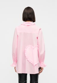 Γυναίκα με κοντά καστανά μαλλιά που φοράει ένα ανοιχτό ροζ μπλούζα με μακρύ μανίκι, με βολάν στις μανσέτες και ένα μεγάλο καρδιακό μπαλώμα με βολάν στην πλάτη.