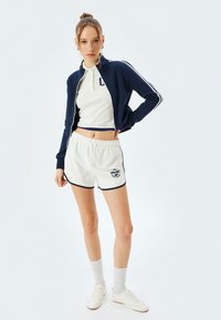 Giacca zip-up blu navy con strisce bianche, top corto a coste bianco, pantaloni corti crema con rifiniture blu navy e grafica. Calzini e scarpe bianche completano il look.