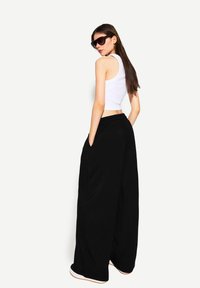 Svarta vida byxor med fickor, i kombination med en vit kortärmad linne. Modellen bär mörka solglasögon, har håret utsläppt och vänder sig något bakåt.