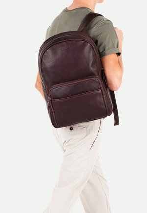 Leather backpack in dark brown with a textured finish, featuring one main compartment and a front zipper pocket, worn on one shoulder.