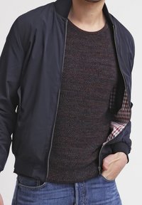 Giacca bomber blu sopra un maglione a maglia scura e testurizzata, con un motivo a scacchi visibile all'interno della giacca, abbinata a jeans blu.