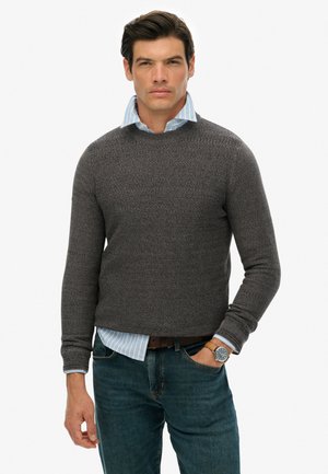 Suéter de punto gris con cuello redondo, tejido texturizado, combinado con una camisa de cuello con rayas azul claro. Llevado con jeans oscuros y un reloj.