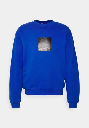Felpa blu con scollatura tonda e maniche lunghe. Una grafica quadrata nera sul petto riporta la scritta "KARL LAGERFELD JEANS."