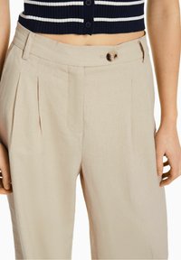 Beige linnebyxor med framvikta veck, en knappstängning framtill och sidofickor. Midjebandet är platt med bälteshällor.