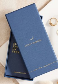 Boîte texturée bleu marine avec un texte doré, "Violet Hamden," et un design de croissant de lune. À l'intérieur, un revêtement bleu marine sans embellissement.