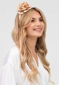 Beżowa opaska do włosów z kwiatem z tkaniny, noszona z długimi falującymi blond włosami, na jasno szarym tle. Widoczna biała odzież.