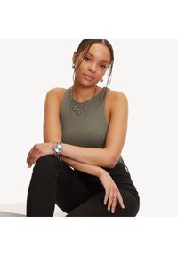 Olijfgroene mouwloze top, zwarte broek, zilveren kettingen sieraden, ronde horloge met schakelarmband, gladde textuur, zittende houding.