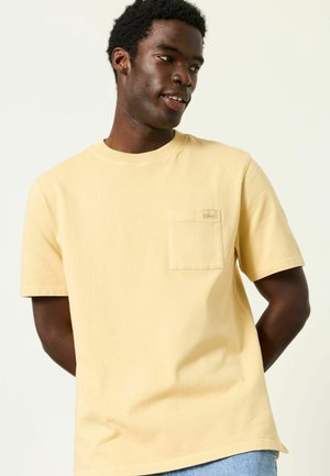 Mann trägt ein hellgelbes T-Shirt mit kurzen Ärmeln und Brusttasche, schaut seitlich, Arme hinter dem Rücken gegen einen schlichten hellen Hintergrund.