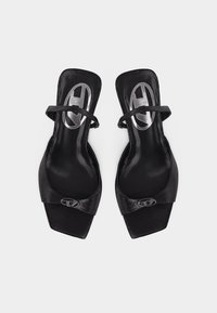 Μαύρα σανδάλια με τετράγωνη μύτη, λεπτά λουράκια αστραγάλου και φαρδιά, υφή μπροστινά λουριά, φωτογραφημένα από πάνω σε απλό φόντο.