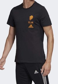 Černé bavlněné tričko s oranžovým emblémem lva a nápisem "HOLLAND" nad logem adidas. Krátké rukávy, design s kulatým výstřihem.