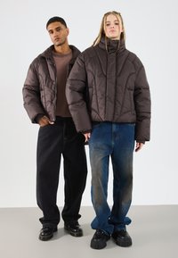 Două modele stau îmbrăcate în jachete puffy maro închis, blugi largi și pantofi negri masivi, pe un fundal alb simplu.