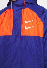 Větruvzdorná bunda Nike s modrým tělem a jasně oranžovými rameny. Má kapuci, přední zip a dvě bílé loga Swoosh na hrudi.
