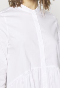 Camicia bianca con bottoni, realizzata in cotone morbido, caratterizzata da un colletto alla coreana e un pannello inferiore arricciato per maggiore texture e dettaglio.