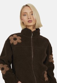 Veste en polaire marron avec un col montant et une fermeture éclair. Détails en appliqué floral de couleur marron clair sur les épaules. Tissu doux et texturé.