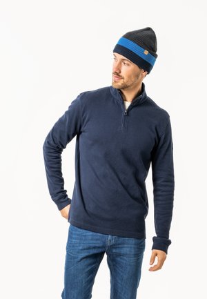 Dunkelblauer Strickpullover mit einem Halb-Reißverschluss-Kragen, kombiniert mit einer dunkelblauen, gestreiften Mütze. Das Model trägt Jeans.